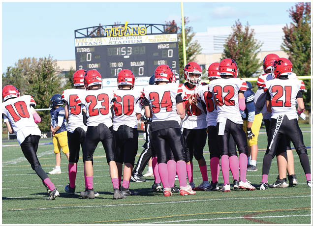 A group of football players in red helmets and white jerseys with pink socks are gathered on a field near a scoreboard showing a tied score. Description generated by AI
