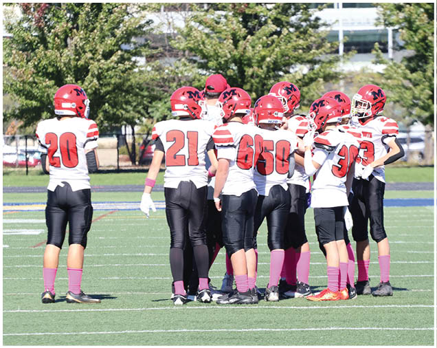 A group of people wearing red helmets, white jerseys with red numbers, black pants, and pink socks are gathered on a football field with trees and buildings in the background. Description generated by AI