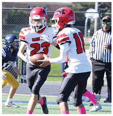 Two people in red and white football uniforms with pink socks are playing on a field while a person in a blue and yellow uniform approaches and two adults stand in the background near a fence. Description generated by AI