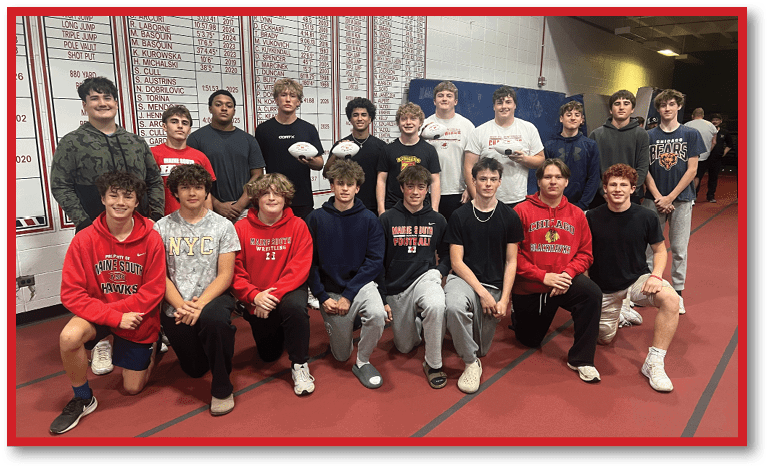 A group of people wearing casual athletic clothing pose together indoors on a track with a scoreboard or record board visible on the wall behind them. Description generated by AI