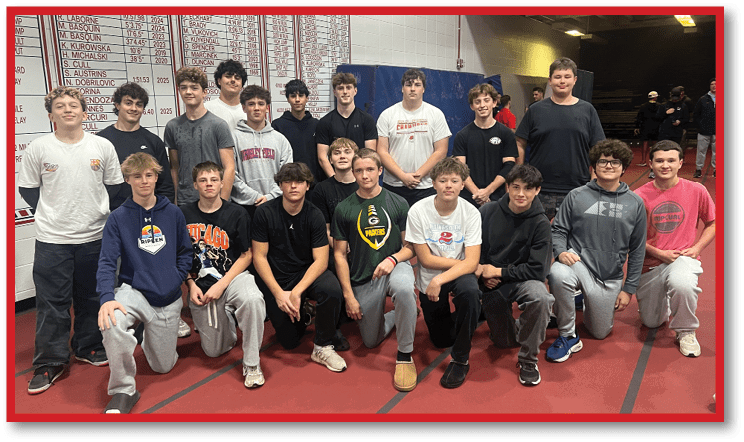 A group of people wearing casual clothing pose together indoors in front of a wall displaying sports records for a high school. Description generated by AI