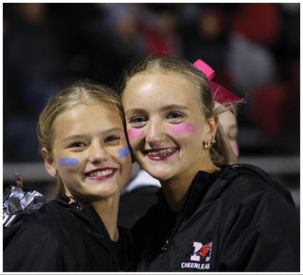 Two smiling people with face paint on their cheeks, one with braces and a pink bow in their hair, posing closely together. Description generated by AI