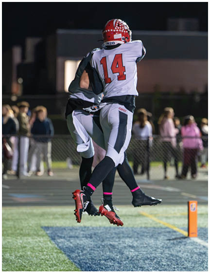 Two athletes in football uniforms jump simultaneously on a field during a game with blurred spectators in the background. Description generated by AI