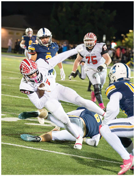 A group of people in football uniforms are actively playing on a field with one person in white falling while holding the ball and others in blue and white uniforms attempting to tackle or block. Description generated by AI
