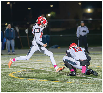 Two people in football uniforms are on a field at night with one person holding the ball on the ground while the other is kicking it. Description generated by AI