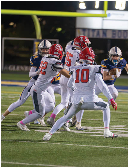 A group of people in football uniforms are actively playing on a field with one person in a dark uniform running with the ball while others in white uniforms attempt to block or tackle. Description generated by AI