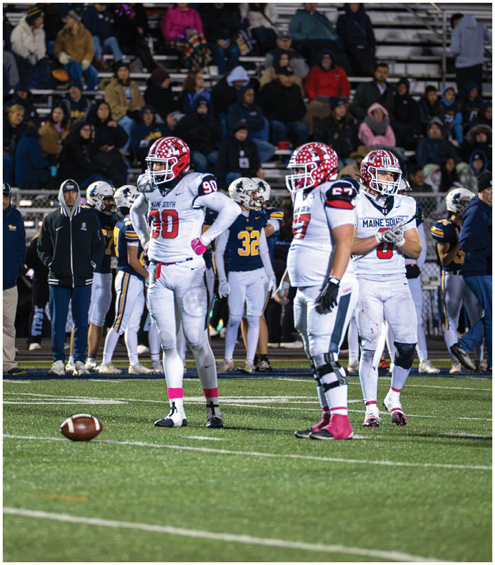 Players in football uniforms stand on a field with a football on the ground while spectators watch from the bleachers in the background. Description generated by AI