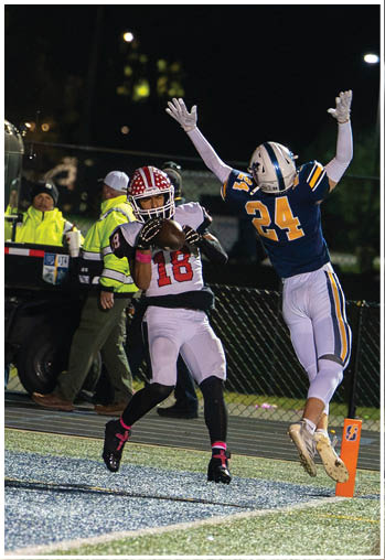 A person in a football uniform with a helmet and long sleeve arm covering is reaching up with one hand while another person in a different football uniform is partially visible. Description generated by AI