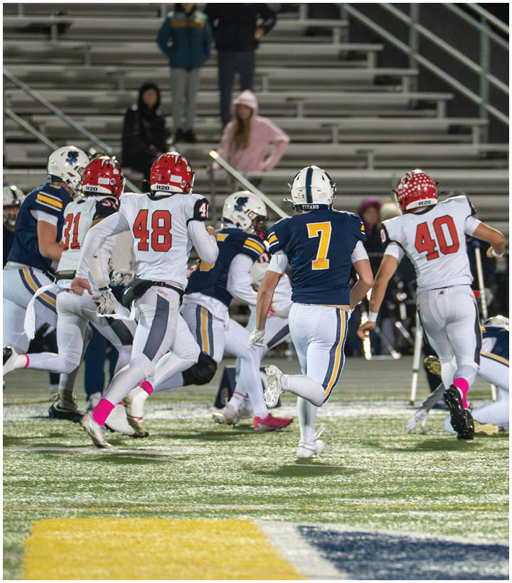 Several people wearing football uniforms are actively running on a field during a game with empty bleachers in the background. Description generated by AI