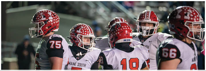 Five people wearing football helmets and uniforms are standing together on a field, appearing to be in conversation or strategizing during a game. Description generated by AI