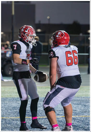 Two people wearing football uniforms and helmets are on a field at night, with one holding a football and the other preparing to receive it. Description generated by AI