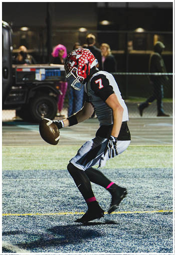 A person wearing a football uniform with a red helmet is holding a football and appears to be preparing to kick it on a lit field at night. Description generated by AI