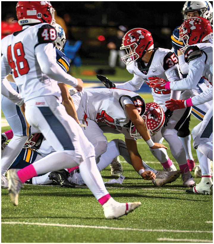 Several people in football uniforms are engaged in a play on a grassy field with one person on the ground and others reaching or moving around them. Description generated by AI