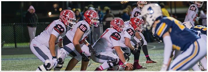 A group of people in football uniforms are lined up on a field in a ready stance, preparing for a play during a game. Description generated by AI