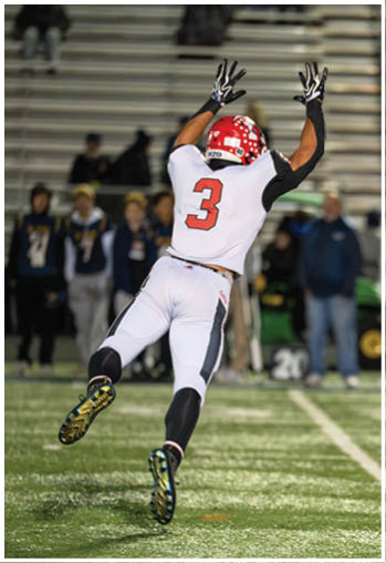 A person wearing a red helmet and white sports uniform with the number three is reaching up with both arms on a football field. Description generated by AI