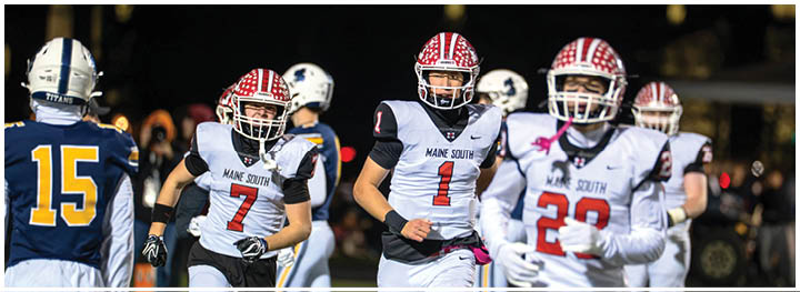 People wearing football uniforms are on a field with some running and others standing, with a crowd visible in the background. Description generated by AI