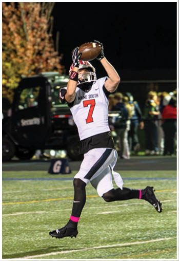 A person wearing a football helmet and a Maine South jersey with the number seven is catching a football during a game. Description generated by AI