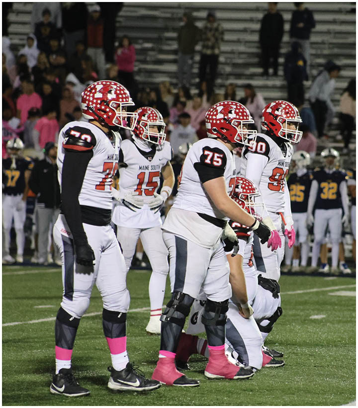 A group of football players in white uniforms with red helmets and pink accessories stand on a field with spectators and other players in the background. Description generated by AI