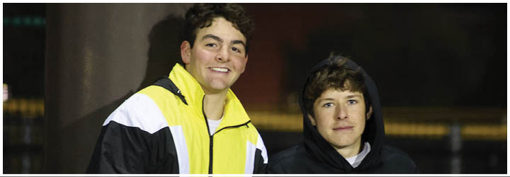 Two people stand close together outdoors at night, one wearing a yellow and white jacket and smiling, while the other wears a dark hoodie and looks forward. Description generated by AI
