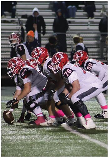 A group of football players in white uniforms with red helmets and pink socks are lined up on a field preparing for a play during a nighttime game. Description generated by AI