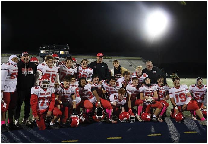 A group of people wearing football uniforms and coaches in team apparel pose together on a football field at night under bright stadium lights. Description generated by AI