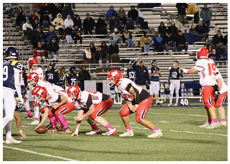 Players in red and white uniforms are lined up on a football field preparing for a play against opponents in dark blue and white uniforms with spectators in the background. Description generated by AI