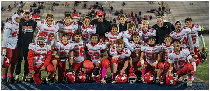 A group of people wearing red and white football uniforms and helmets pose together on a football field with empty bleachers in the background. Description generated by AI