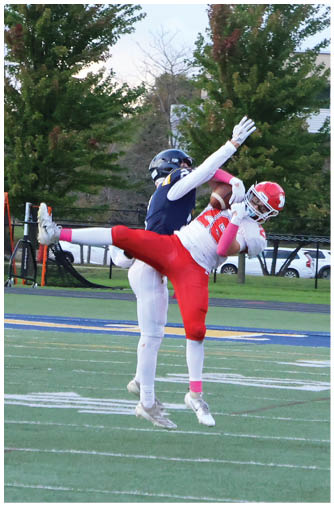 Two people wearing football uniforms are engaged in a play on a field with one person in red pants and a white jersey leaning backward while the other in white pants and a dark jersey reaches over them. Description generated by AI