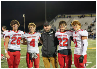 Four people in football uniforms and one person in casual outdoor clothing stand together on a football field at night with stadium seating in the background. Description generated by AI
