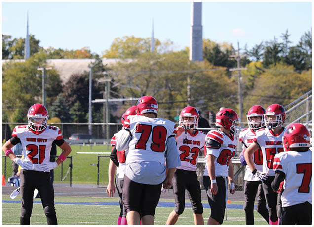 A group of people wearing red helmets and white sports jerseys are standing on a football field with bleachers and trees in the background. Description generated by AI