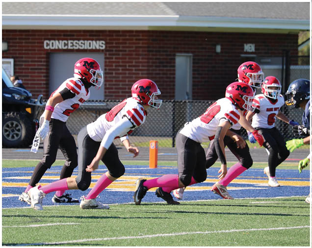 Several people in football uniforms with red helmets and pink socks are lined up on a field facing opponents in dark helmets and yellow pants during a game. Description generated by AI