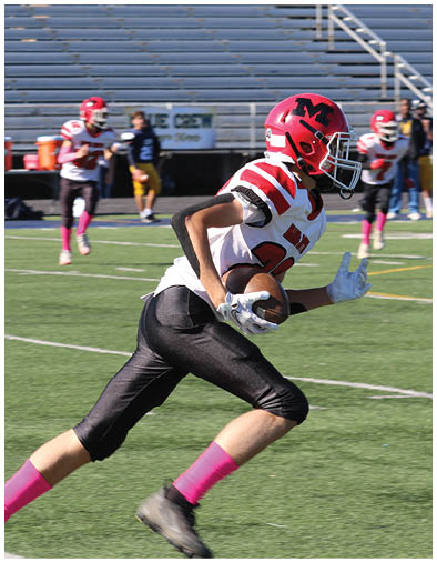 A person wearing a red helmet and white jersey is running on a football field with other players and people standing in the background near empty bleachers. Description generated by AI