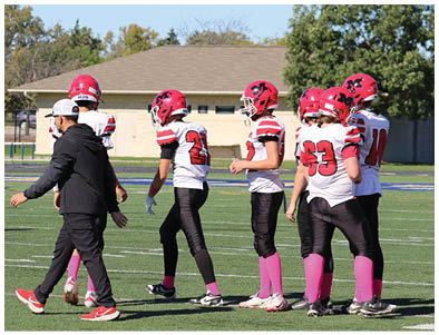 A group of people wearing red helmets, white jerseys with black and red stripes, black pants, and pink socks are standing and walking on a football field with a coach in black sportswear. Description generated by AI