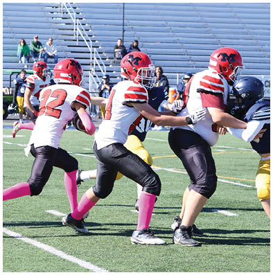 Three people in red helmets and white jerseys with pink socks are playing football on a field with empty bleachers in the background. Description generated by AI