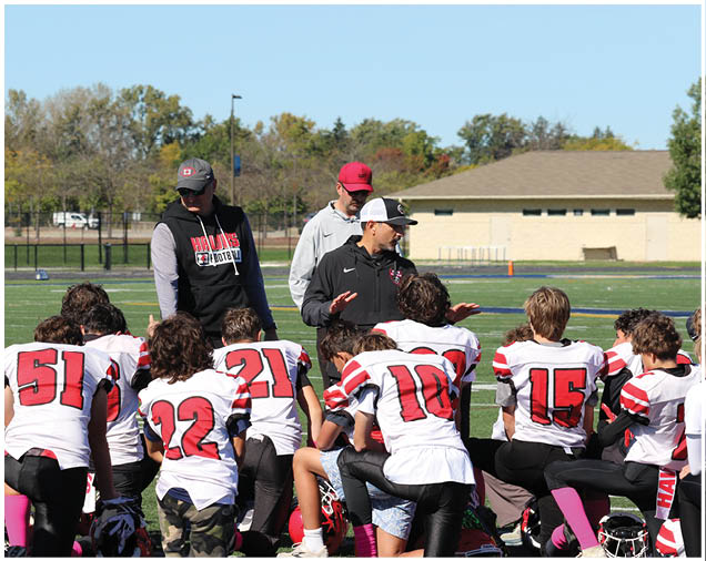 A group of people wearing football uniforms with numbers on their backs are kneeling on a field while three adults in casual sportswear stand and talk to them. Description generated by AI