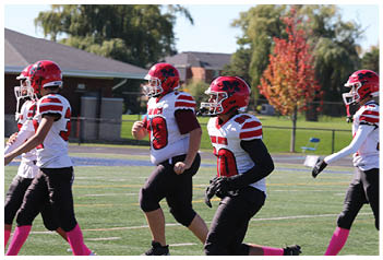 Four people wearing red helmets, white jerseys with red stripes, black pants, and pink socks are walking on a football field with trees and buildings in the background. Description generated by AI