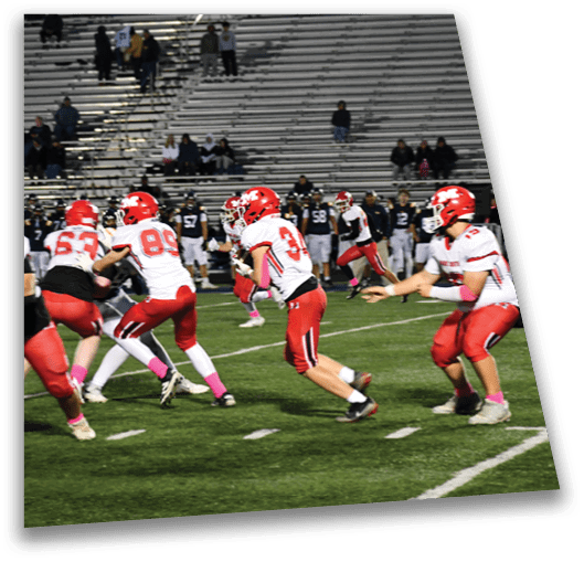 People in red and white football uniforms are actively playing on a green field with empty bleachers in the background. Description generated by AI