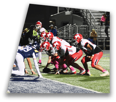 Players in red and white uniforms are lined up on a football field facing players in blue and white uniforms, preparing for a play during a nighttime game with spectators in the stands. Description generated by AI