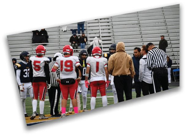 People in football uniforms and coaches are gathered on a field with empty bleachers in the background. Description generated by AI