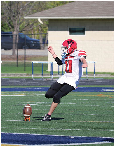 A person wearing a red helmet and black athletic gear is preparing to kick a football placed on a tee on a green field with hurdles and a fence in the background. Description generated by AI