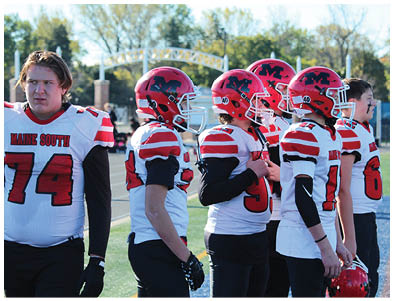 Three people wearing red and white football uniforms and helmets stand on a field, with one person adjusting their jersey. Description generated by AI