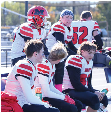 A group of people wearing white and red football uniforms are sitting and standing on a bench outdoors, with one person wearing a red helmet and another wearing a star-spangled bandana. Description generated by AI