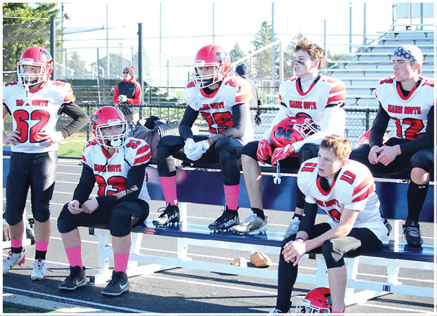 Five people wearing red and white football uniforms with pink socks are sitting and standing on blue bleachers at an outdoor sports field. Description generated by AI