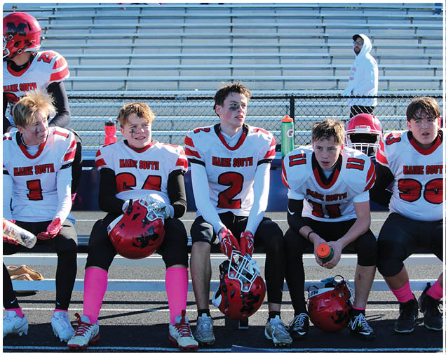 Five people wearing football uniforms with helmets sit on a bench on a sports field with empty bleachers in the background. Description generated by AI