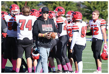An adult wearing sunglasses and a black hoodie holding a football stands near a group of people in red helmets and white football uniforms on a field. Description generated by AI