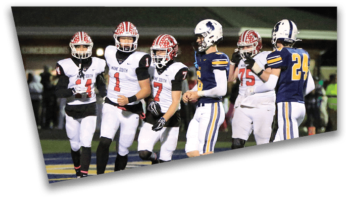 A referee is throwing a football towards three players in football uniforms standing on a field at night with spectators in the background. Description generated by AI