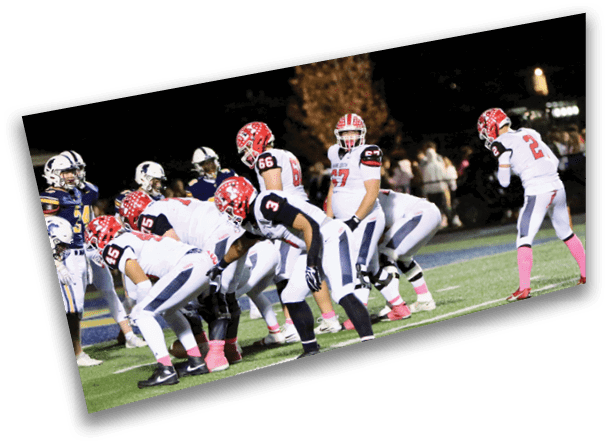 A group of people wearing football uniforms with red helmets and pink socks are lined up on a field at night, preparing for a play against an opposing team in dark uniforms. Description generated by AI