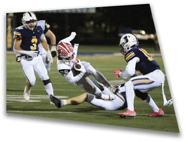 A person in white sports gear with a red helmet is falling while holding a football as two people in blue and white uniforms approach on a football field. Description generated by AI