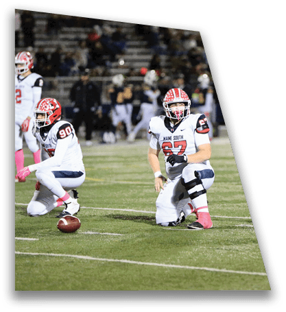 A person wearing a football uniform with a red and white helmet is kneeling on a grassy field during a game. Description generated by AI
