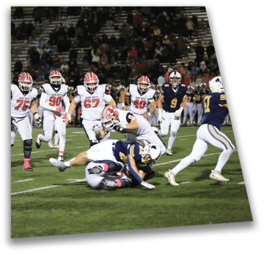 A group of people wearing football uniforms are actively playing on a field with one person tackling another who is holding the ball. Description generated by AI